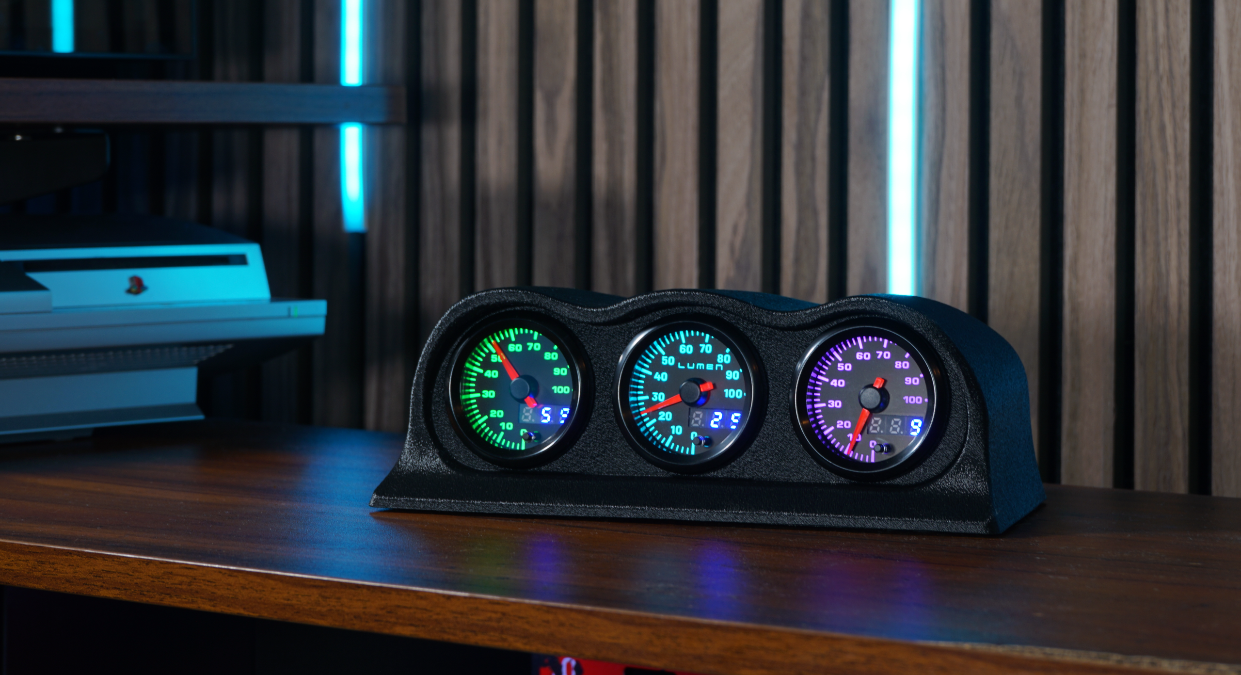 The Lumen Dash three-gauge PC hardware monitor on a wooden desk, illuminated in green, blue, and purple.
